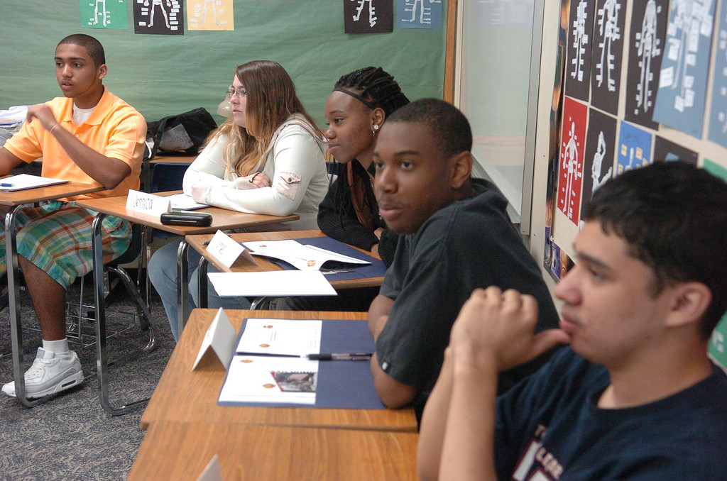 A diverse group of students sitting at desks looking intently to something out of frame.