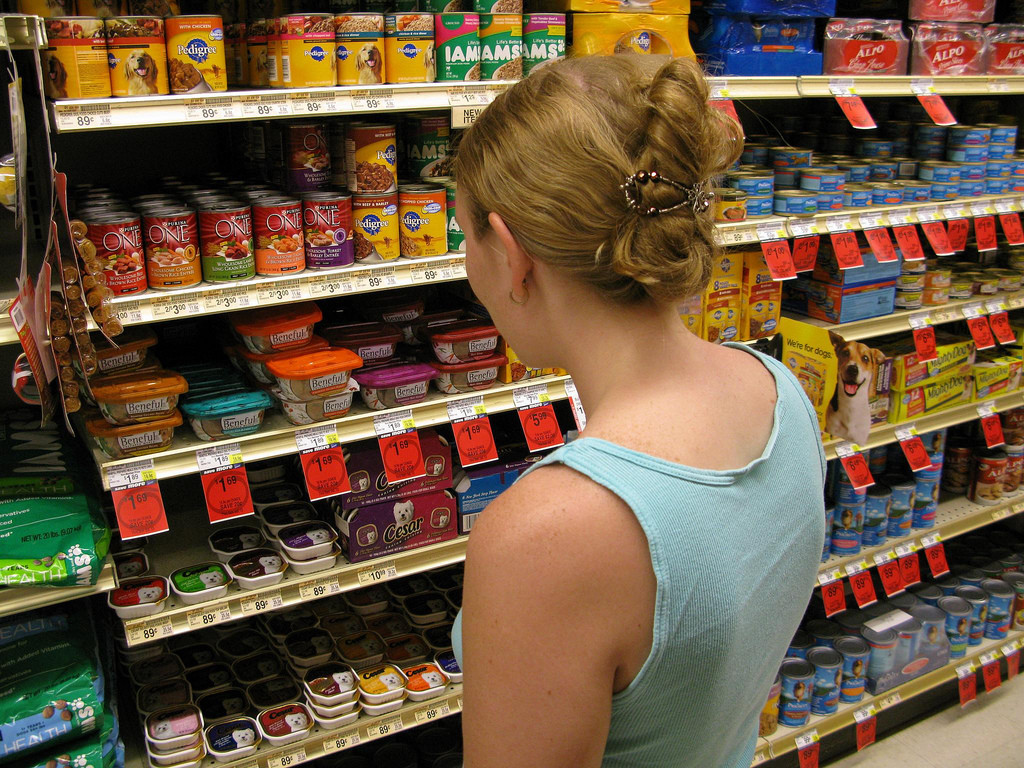 Vista de la mujer desde atrás, de pie frente a los estantes de las tiendas de comestibles, mirando la selección de alimentos para perros.