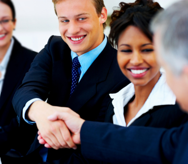 A man in a suit smiling and shaking hands with another man. A woman in a suit is in between them, also smiling.