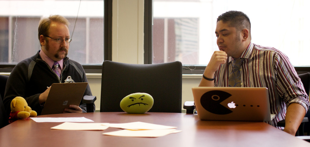 Two men are seated in a meeting room; both look frustrated. In between them is a chair for the boss, who is absent, but is represented by a small green pillow on the table with a "frowny face."