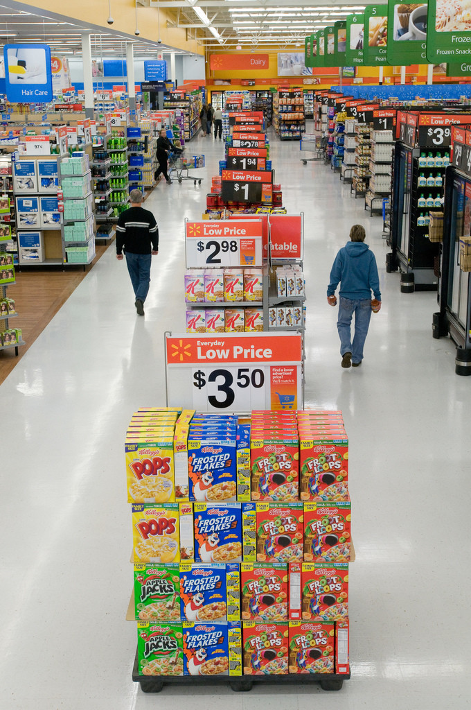 Foto van een winkelgang in Walmart. Op de voorgrond staan in het midden van het gangpad grote displays met ontbijtgranen. Op de achtergrond staan drie shoppers.