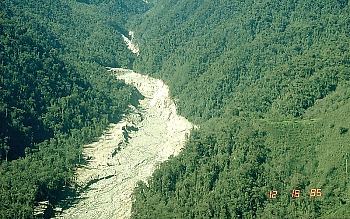 Rio que flui através de uma floresta; o rio é misturado com fragmentos de rocha, fazendo a água parecer cinzenta e grossa.