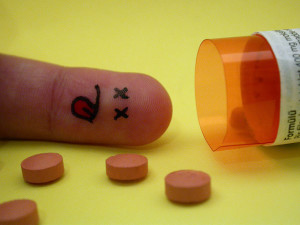 Finger with a dead face drawn on it, next to a spilled pill bottle
