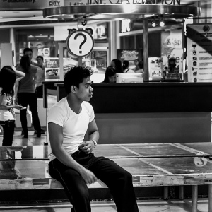 Black and white photo of a young man sitting in front of a City Information booth, so that the question mark sign is positioned directly over his head like a thought balloon.