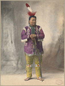 Colorized photograph of a Native American man, standing, propping his hands on a rifle that is pointed towards the floor. He wears a large red and white feather in his hair, a purple shirt with white fringe, yellow pants with a beaded design on the knees, and beaded moccasins.