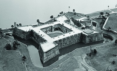 Zdjęcie lotnicze ukazuje hiszpański fort Castillo de San Marcos, kwadratową, wysokościenną strukturę zwróconą w stronę wody i zawierającą otaczającą fosę.