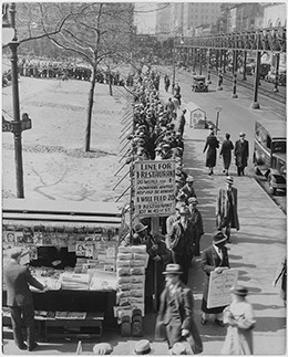 A photograph shows a long line of men waiting on a New York City street for a hot meal. The man at the front of the line holds up a sign that reads, 