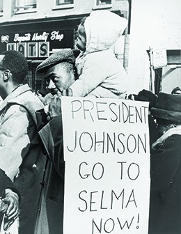 A photograph shows a group of African Americans marching on the street in Selma, Alabama. In the foreground, a man with a small child on his shoulders carries a sign that reads 