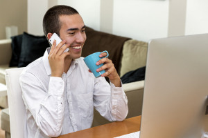 A man using a phone and drinking coffee by a computer.