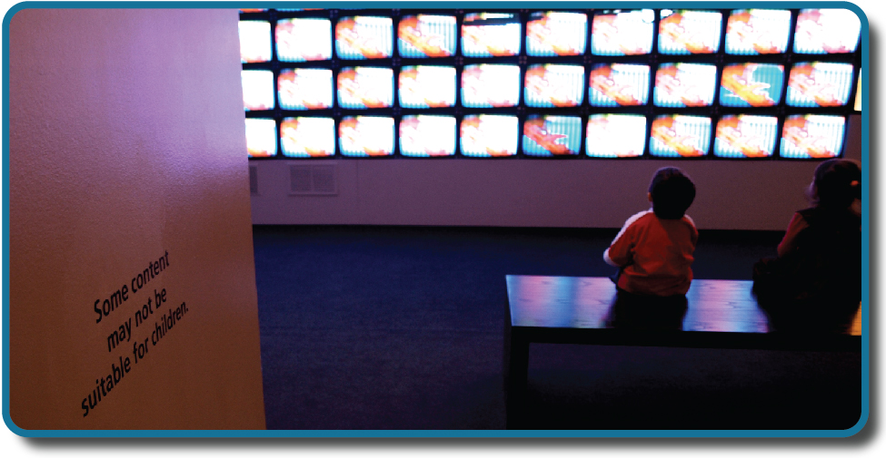 Children sit in front of a bank of television screens. A sign on the wall says, “Some content may not be suitable for children.”