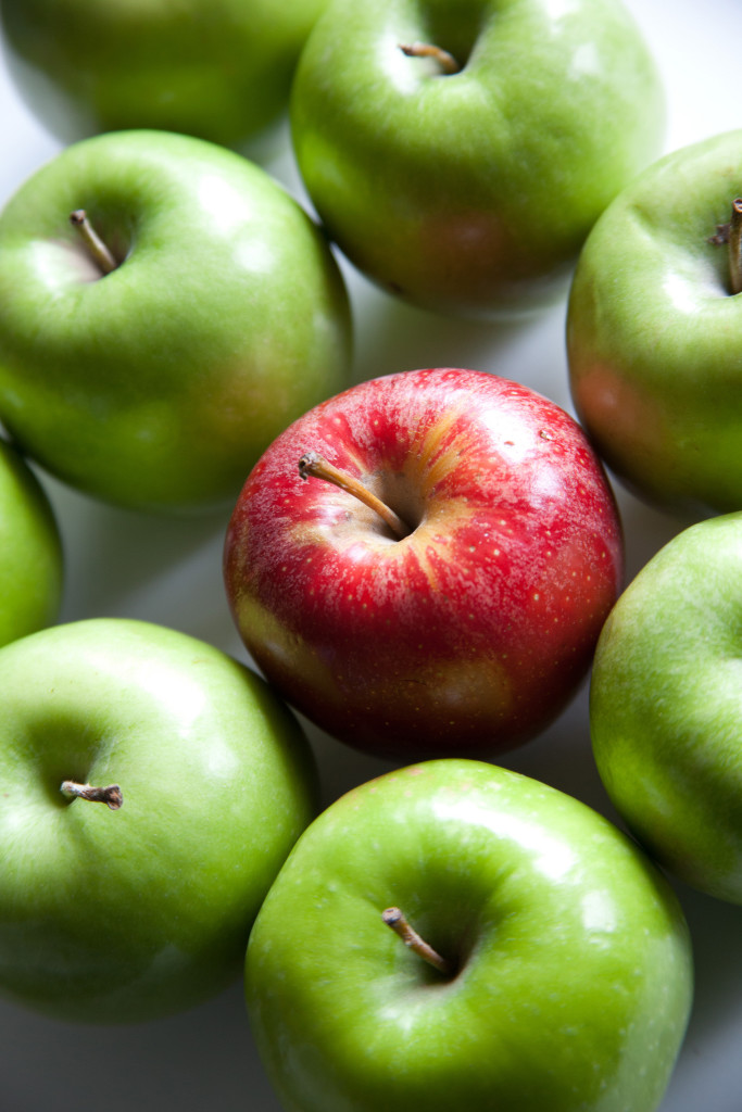 Photo of seven green apples surrounding one red apple.