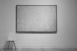 Black and white photo of framed canvas on wall with texture but no image. A white chair sits at bottom left against wall. 