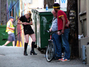 A woman injecting in her arm while two other men stand nearby.