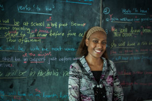 Photo of a female teacher smiling in front of a chalkboard covered with writing.