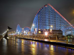 Photo shows a roller coaster at night with lights shining on it, tallest hill is at the forefront of the photo.