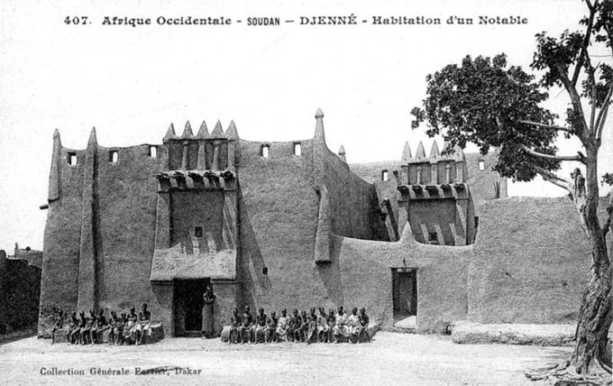 Black and white photo of a Toucouleur-style house with a row of people sitting in front of it.