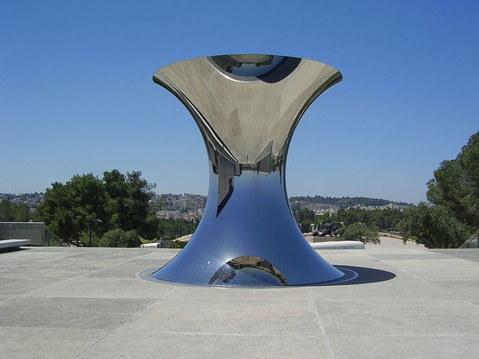 A very large hour-glass shaped object located in a plaza. The shape of the sculpture turns objects that it is reflecting upside down.