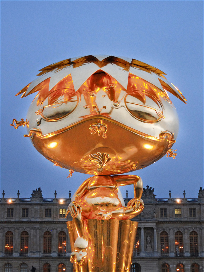 This golden, cartoonish figure has a large head that dwarfs its body. It wears a spiky, flat hat and sits with one leg tucked underneath and one leg dangling down.
