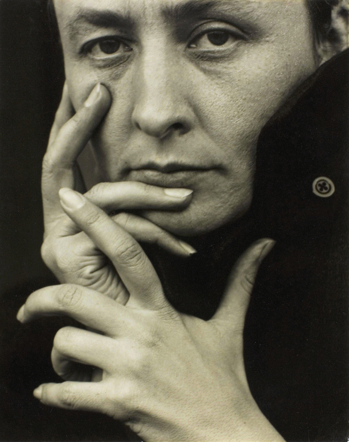 A close-up photo of Georgia O’Keefe with her hands up to her face.