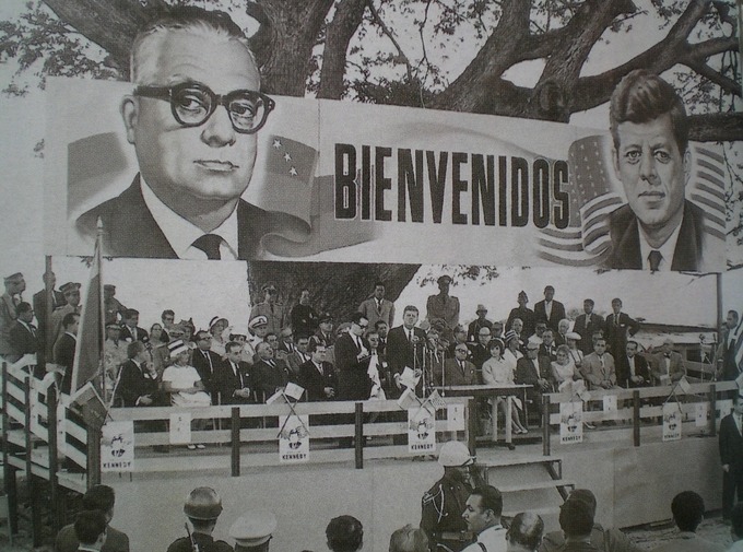 A large billboard over the stage reads "Bienvenidos" and is bookended with portraits of John F. Kennedy and Rómulo Betancourt.