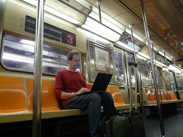 A picture of a man using his laptop while he's riding on a subway train.