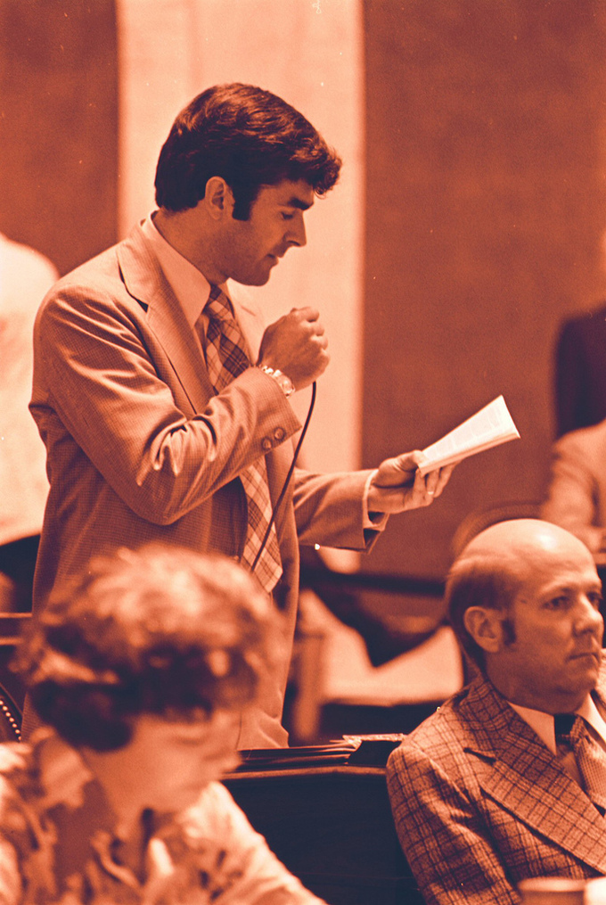 A man holds a microphone and looks at a piece of paper while giving a speech.