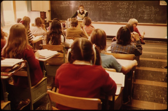 A teacher speaks at the front of a classroom.
