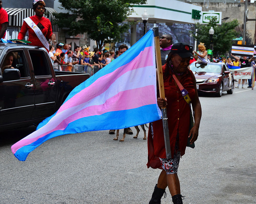 Woman in a parade holding a transgender flag with five horizontal stripes: blue, pink, white, pink, blue.