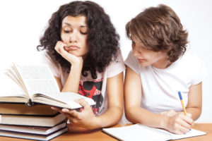 One woman reads a book and another woman, beside her, takes notes.