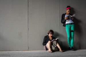 Two women reading. One is standing and leaning against a wall; the other is sitting on the ground, also leaning against the wall.