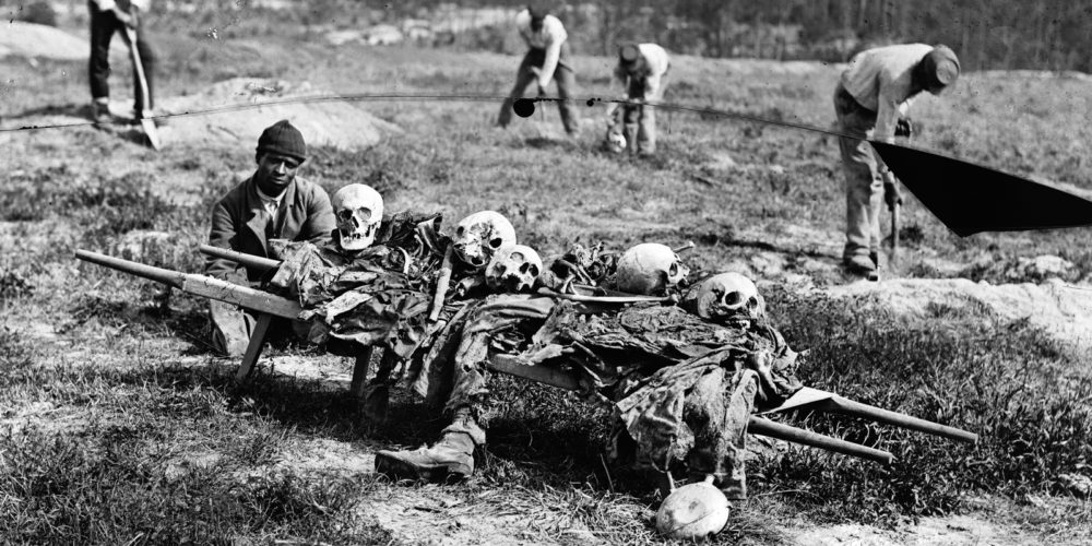 A gruesome photograph, taken after the battle of Cold Harbor, shows a black man squatting next to the skeletal remains of five or six corpses before burial.