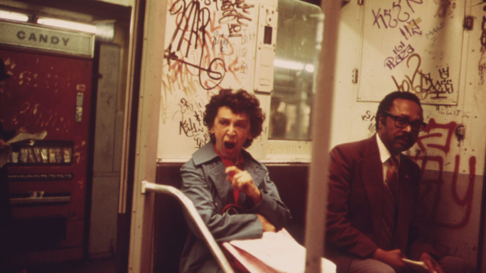 White woman yawns on the subway, sitting next to a black man. The inside of the car is covered in graffiti.