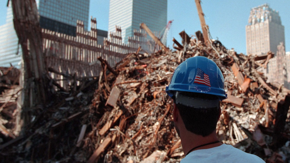 The back of a man's head in a construction hat (with an American flag log) who looks into the massive pile of rubble from the 9/11 attacks.