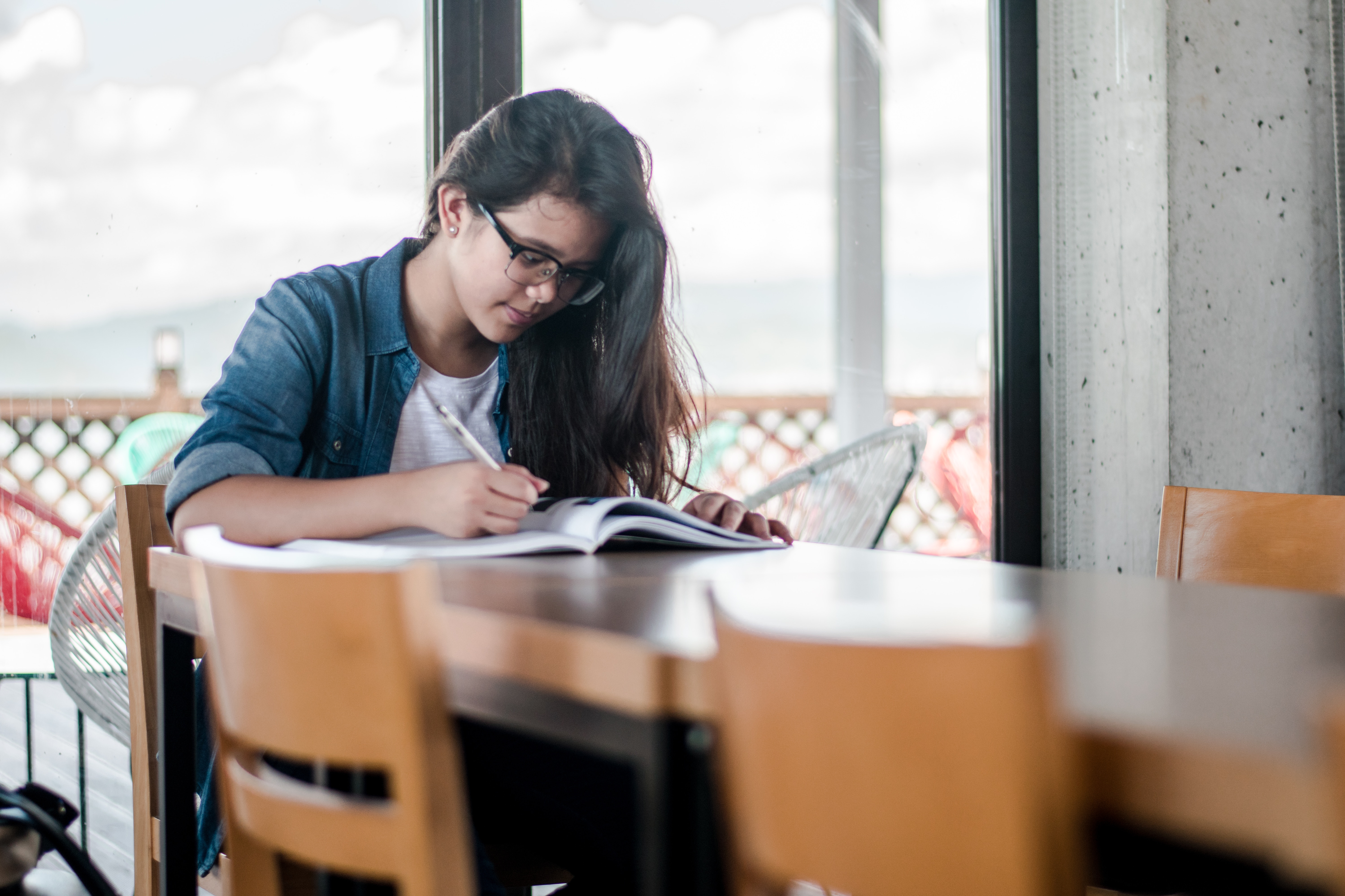 Girl sitting at a long table wearing glasses, writing in an open book.