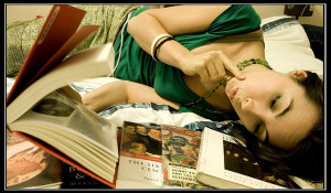 Photo of a woman lying on her side reading a book, with a pile of other books near her head.