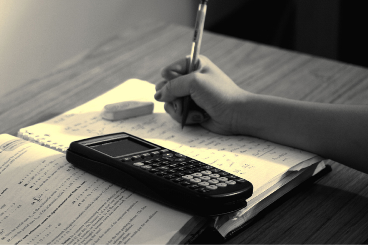 A photograph of a student’s hand, working on a problem with an open textbook, a calculator, and an eraser.