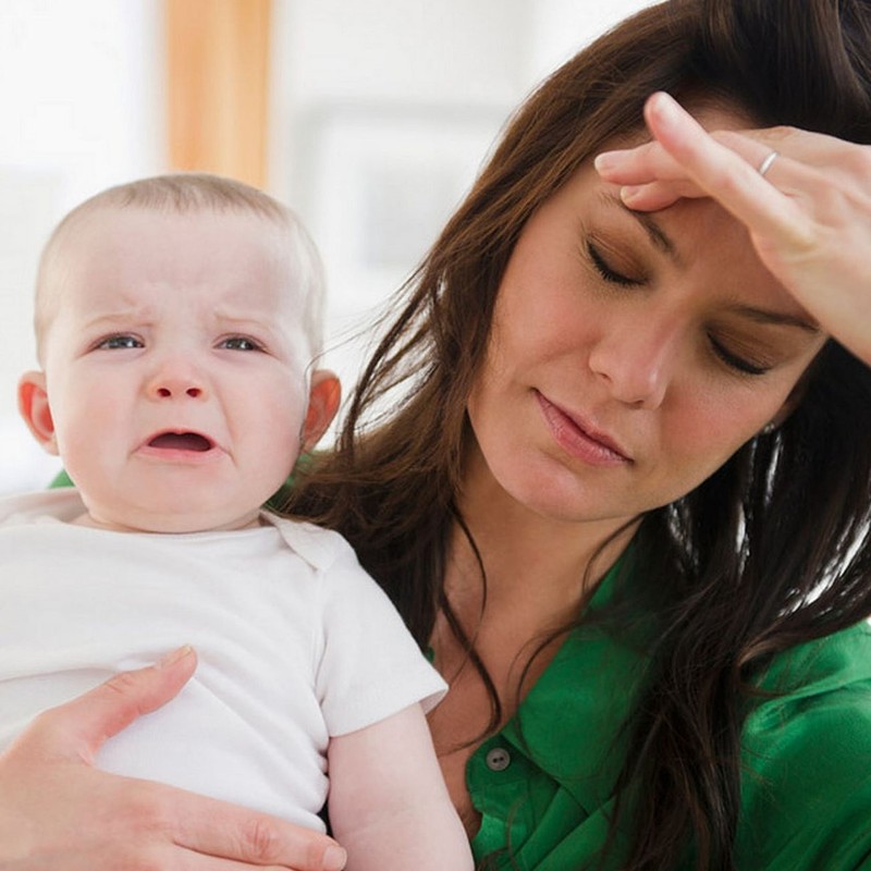 A tired looking mother closes her eyes and rubs her forehead as her baby cries. 