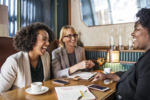 Three women are wearing business attire and sitting at a cafe table drinking coffee and laughing together.