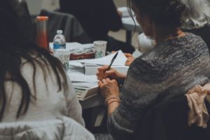 Two women are sitting at a desk with their backs to the camera. One of the women is writing in a book with a pencil. 