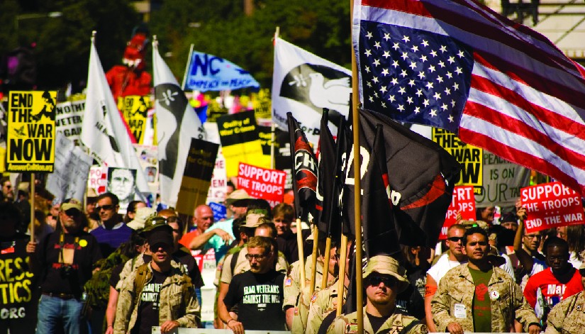 An image of a group of people, several of whom are holding flags and signs. One of the signs reads