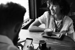 A man and a woman at a coffee shop. The man is looking at his phone, the woman is looking out the window. 