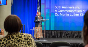 Speaker speaking in front of a projection that reads: 50th Anniversary: A Commemoration of Dr Martin Luther King Jr