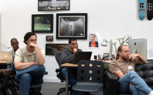 Three men sitting at desks touching their faces and looking in different directions