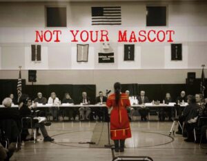 Maulian Dana stands before a school board meeting, the words "Not Your Mascot" appear above her.