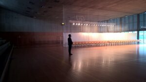 A man standing alone in an empty gym in front of a row of chairs