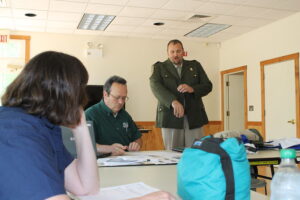 A man speaking to several people wearing a green uniform jacket