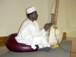 A man in traditional West African clothing sitting on a cushion and playing a string instrument