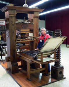 Photograph of a man standing behind a wooden printing press