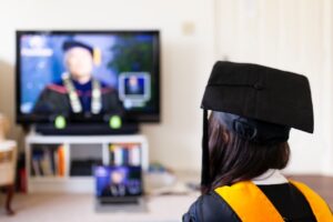 A woman in graduation regalia watches a graduation speech online