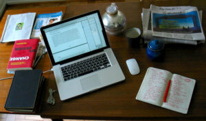 A desk with a computer, notebook, newspapers, books, and papers